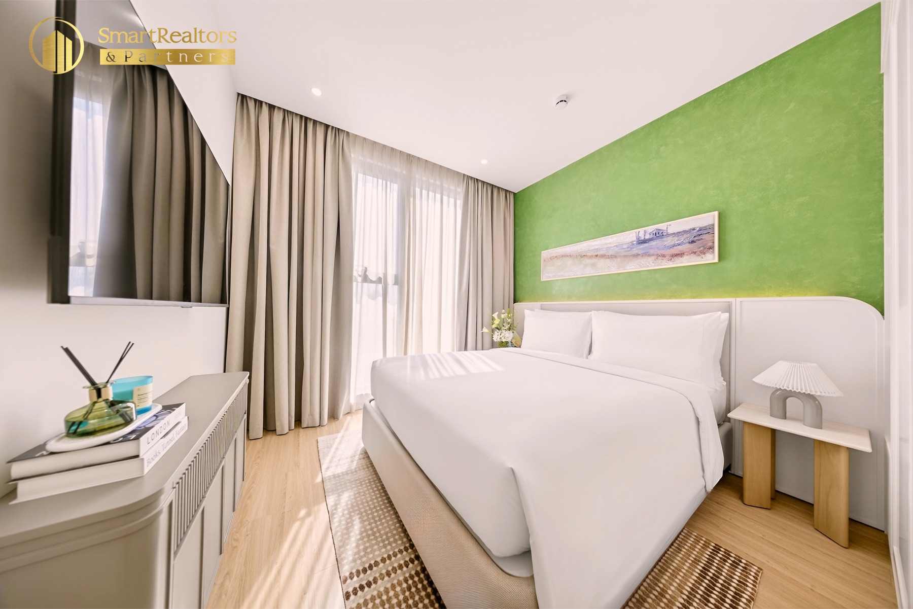 Phòng ngủ căn hộ mẫu 1PN: giường đôi, mảng tường xanh, tranh ngang và tivi treo tường