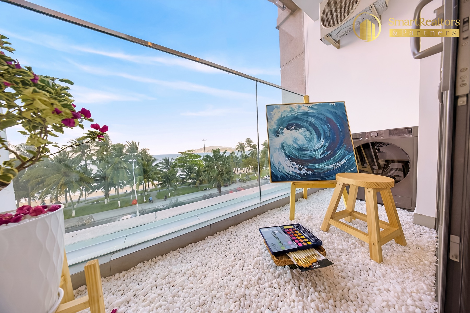 Ban công căn hộ mẫu Studio rải sỏi trắng, giá vẽ tranh và máy giặt, nhìn ra hàng dừa ven biển